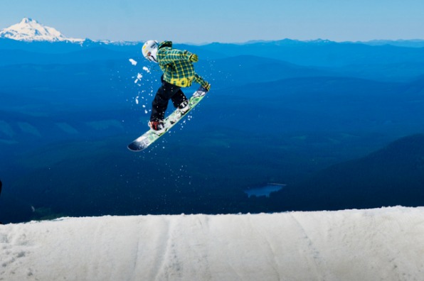 A student snowboarder is seen mid-jump on the side of Mt Hood. In the distance is another snow capped mountain, forested hills and a lake below