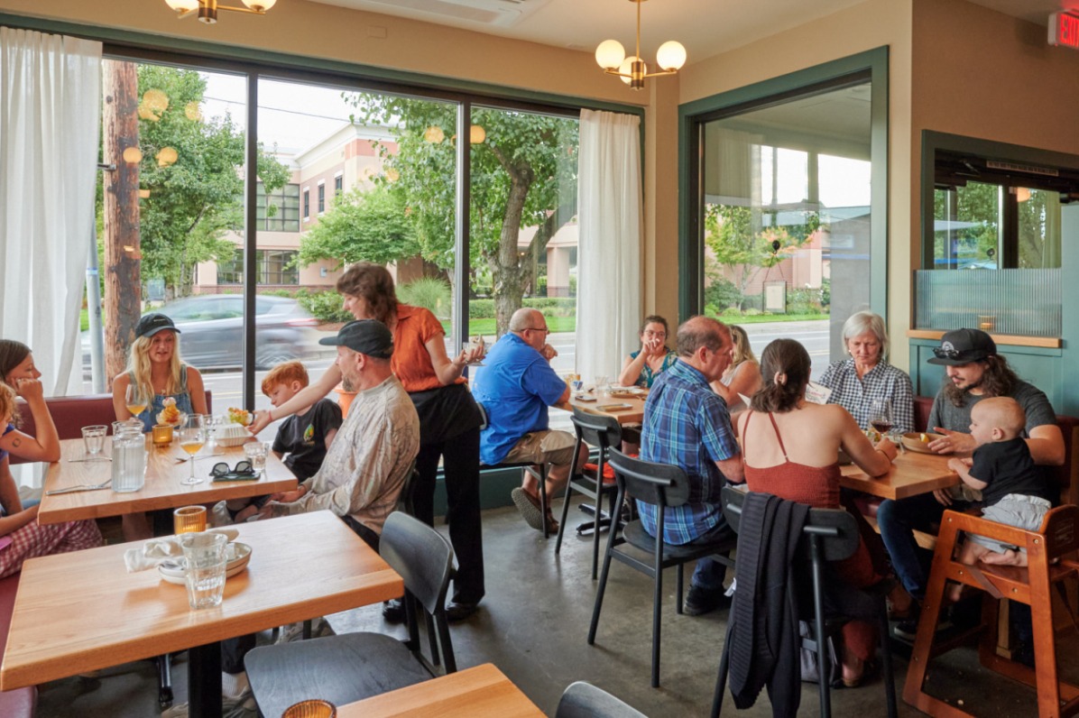 A server at passes out drinks to a table of customers at Canard Restaurant. The dining room is brightly lit and surrounded by windows overlooking a tree lined street. 