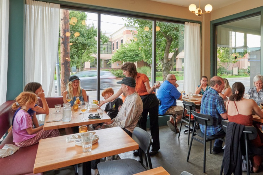 A server at passes out drinks to a table of customers at Canard Restaurant. The dining room is brightly lit and surrounded by windows overlooking a tree lined street. 