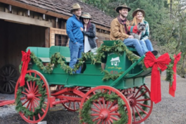 Four people pose for a photo in an antique sleigh. The sleigh is green with red wheels and decorated in holiday greenery and bows. 