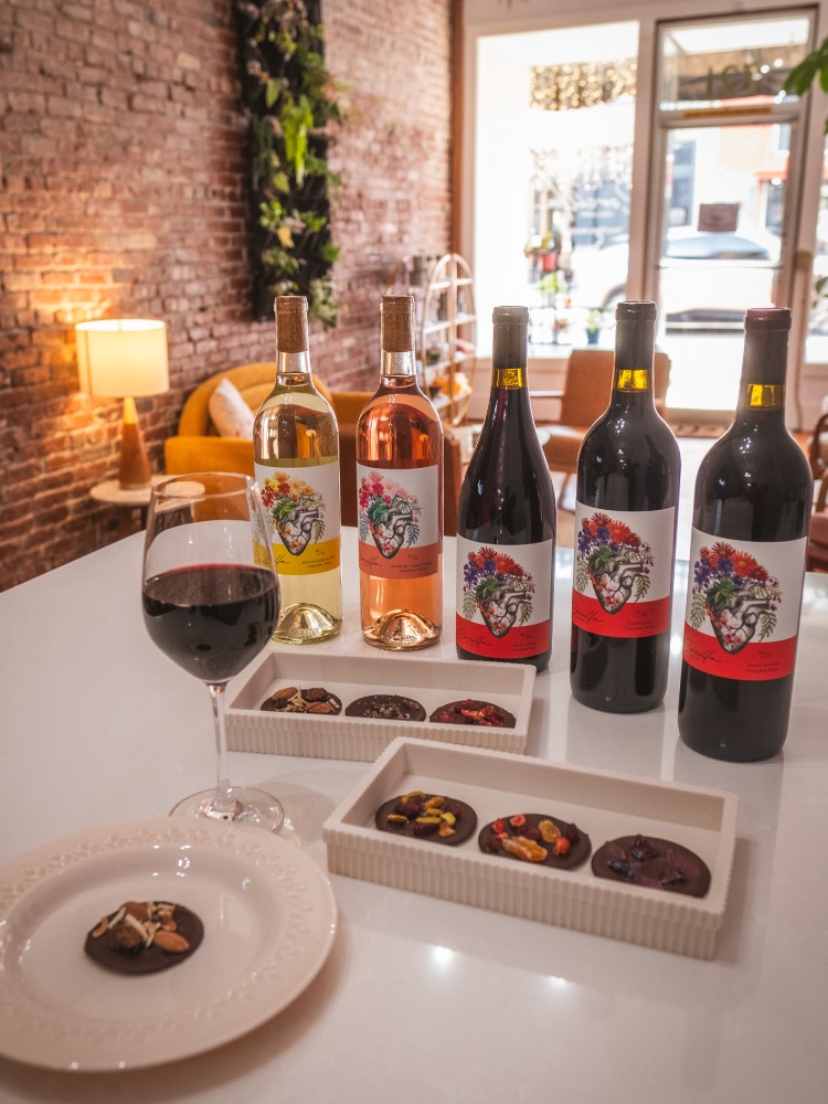 Five bottles of Corcilla Cellars Wines sit on a table with trays of cookies. A half full glass of red wine sits in the foreground. 