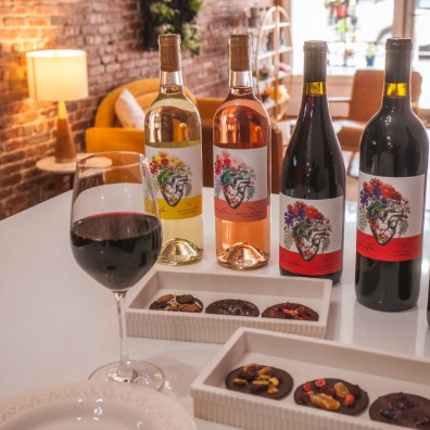 Five bottles of Corcilla Cellars Wines sit on a table with trays of cookies. A half full glass of red wine sits in the foreground. 