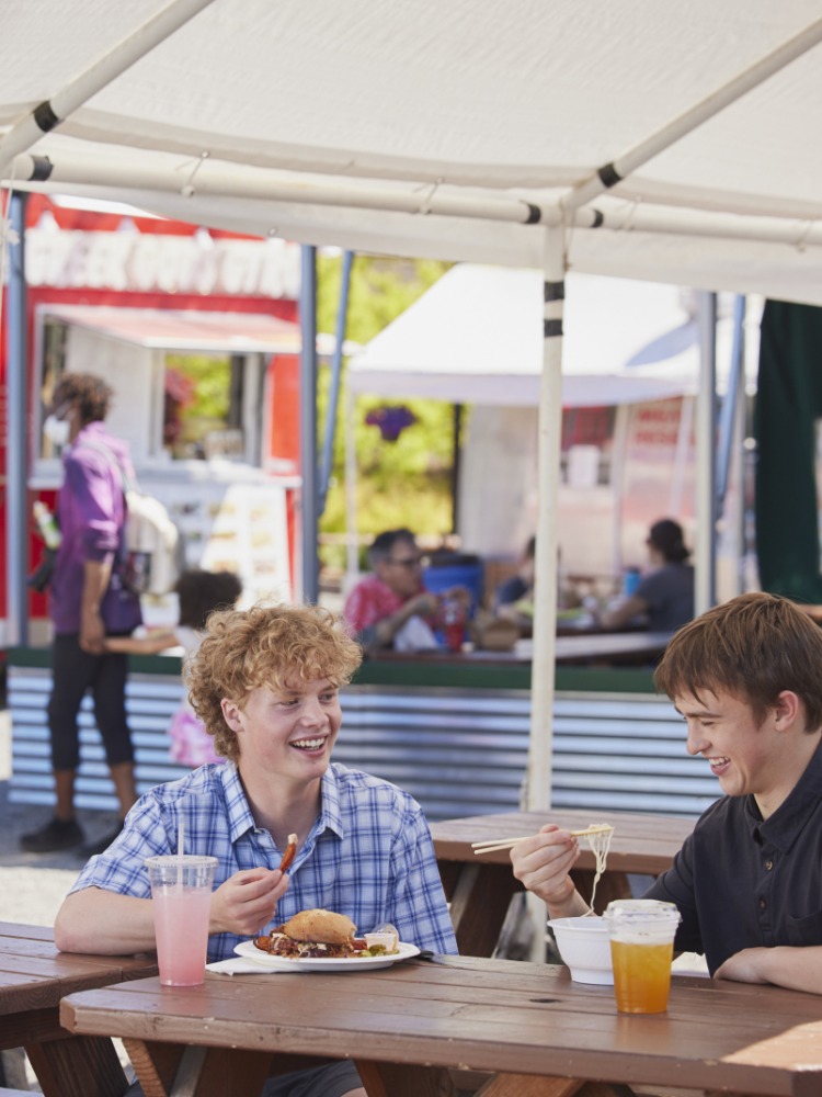 Two people in collared shirts eating at a picnic table with food carts in background