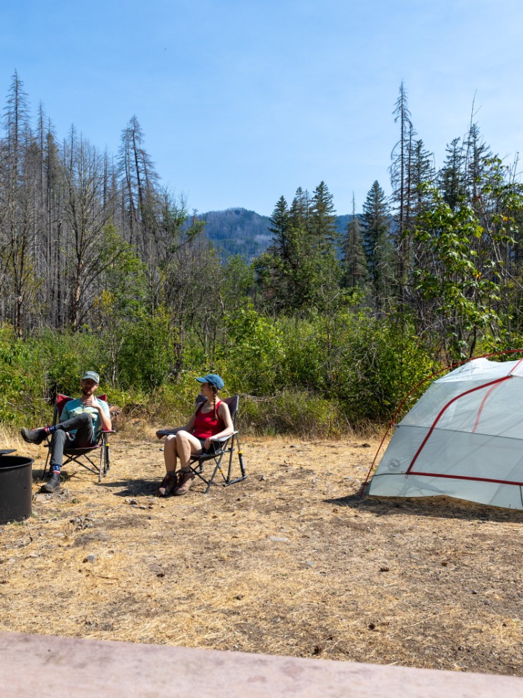 Two campers sit near a fire ring and a tent