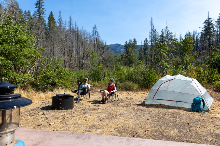 Two campers sit near a fire ring and a tent