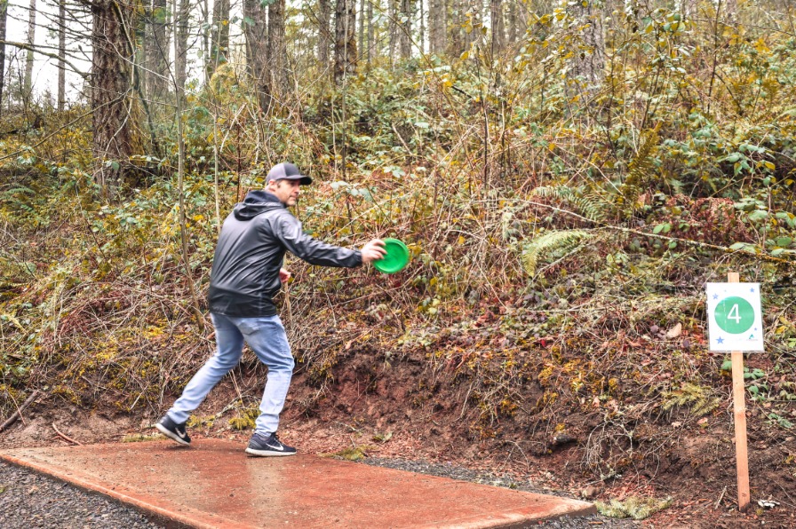 A disc golfers throws a green disc with trees and bushes behind them.