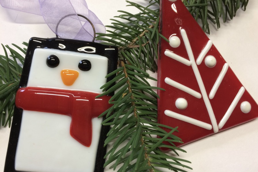 Fused glass ornaments of a a penguin wearing a red scarf and red christmas tree lay atop a holiday tree branch