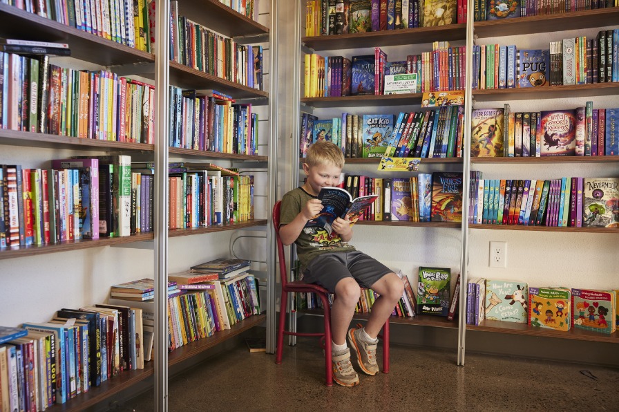 A child reads a book sitting in a red chair surrounded by shelves filled with more books.