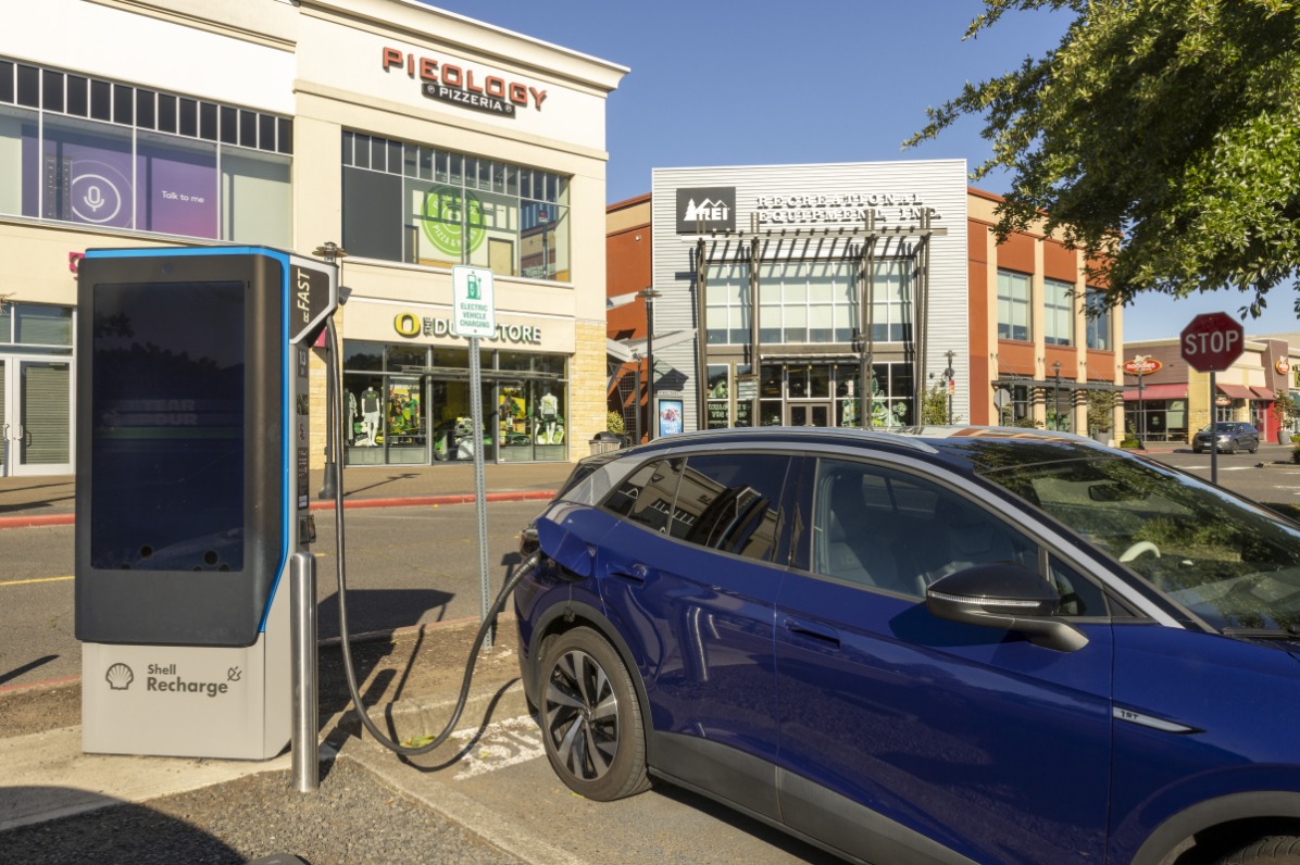 A blue car is plugged into an electronic vehicle charging station with stores behind.