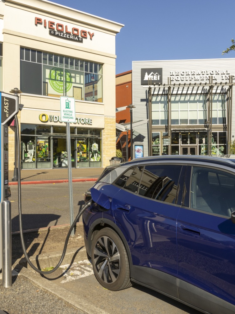A blue car is plugged into an electronic vehicle charging station with stores behind.