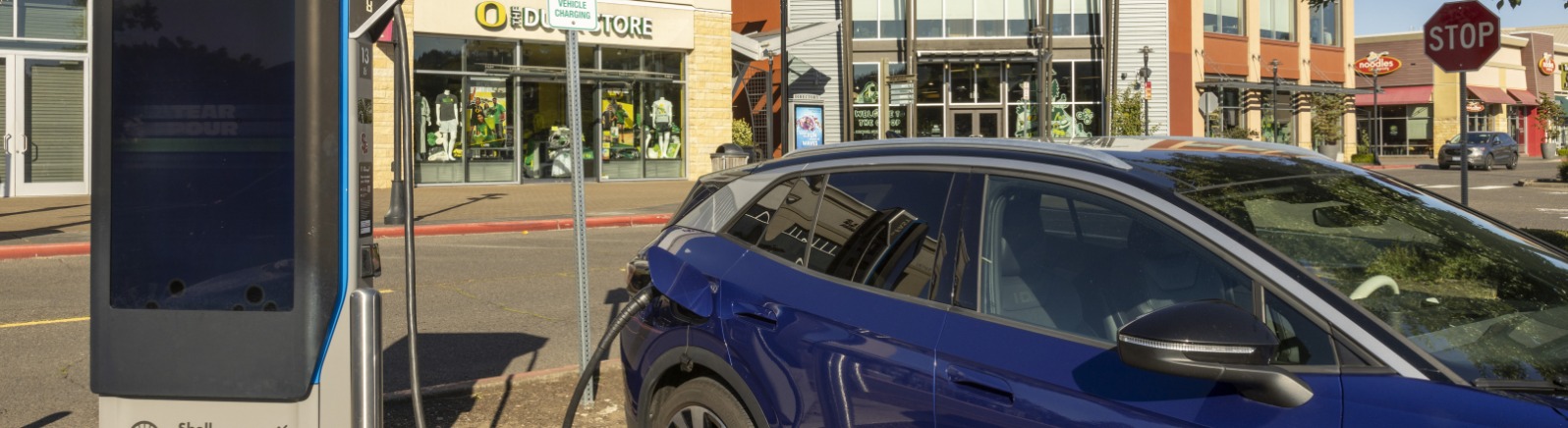A blue car is plugged into an electronic vehicle charging station with stores behind.