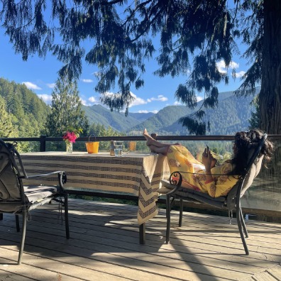 A visitor lounges in a chair with their feet kicked up on the table on the patio of Forest Retreat. The background view is layers of tree lined hills.