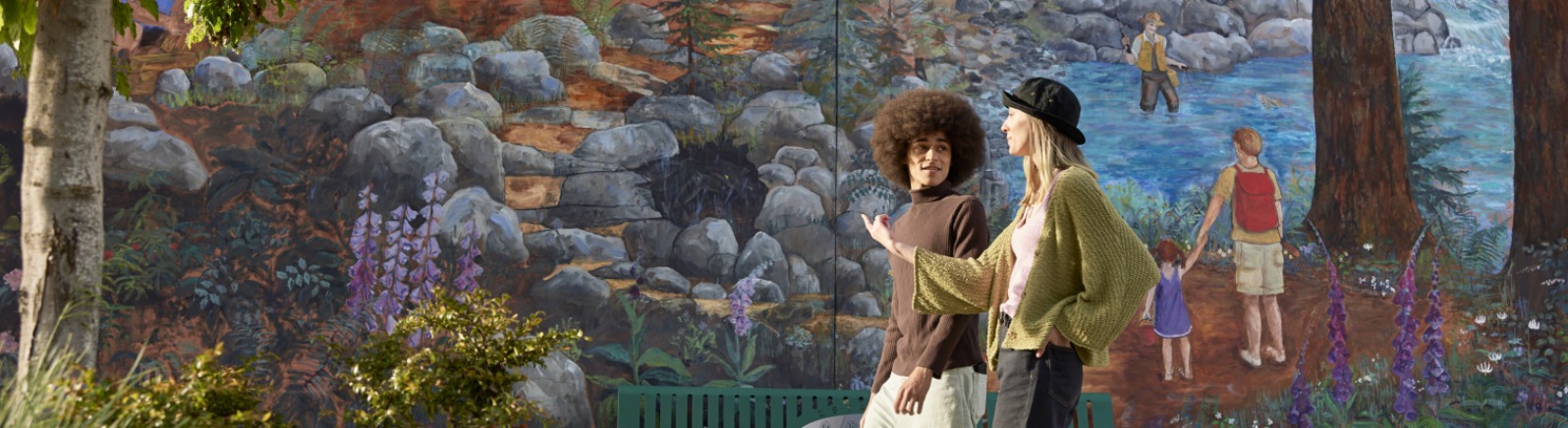 Two visitors walk along a grey sidewalk with a brightly colored mural of a river behind them