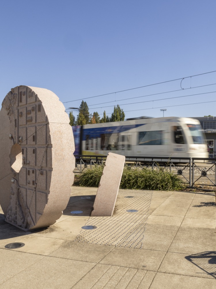 A tan giant circle statue sits in grey cement with a blurry Max train rushing behind it.