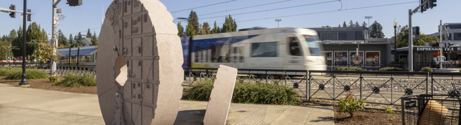 A tan giant circle statue sits in grey cement with a blurry Max train rushing behind it.