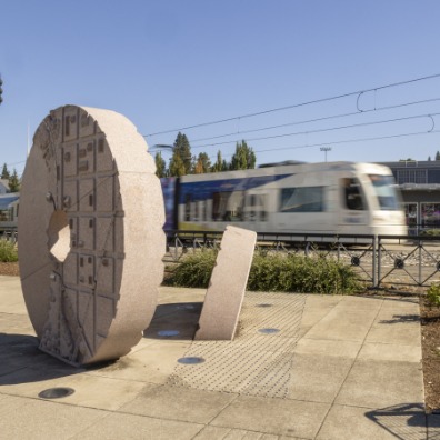 A tan giant circle statue sits in grey cement with a blurry Max train rushing behind it.