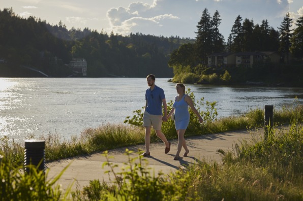 Two walkers along a cement path with green grass on each side and water behind them.