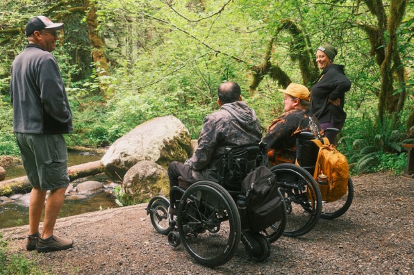 Hikers in wheelchairs and on foot converse at the side of a creek. 