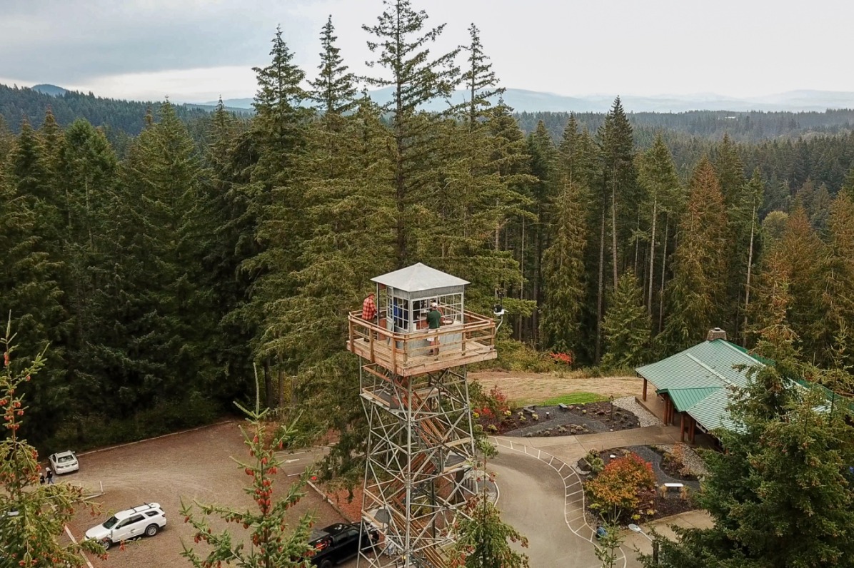 Drone view looking down at fire lookout tower with green evergreen trees behind.