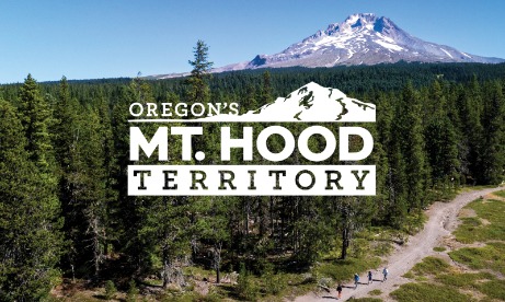 Logo for Oregon's Mt. Hood Territory overlayed on a drone image of Mt Hood, the forest and hikers on a trail.