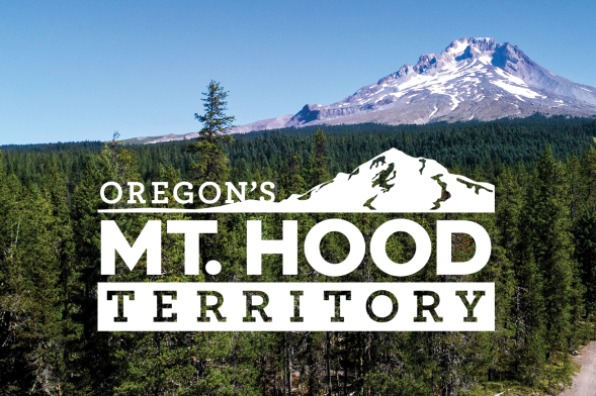 Logo for Oregon's Mt. Hood Territory overlayed on a drone image of Mt Hood, the forest and hikers on a trail.
