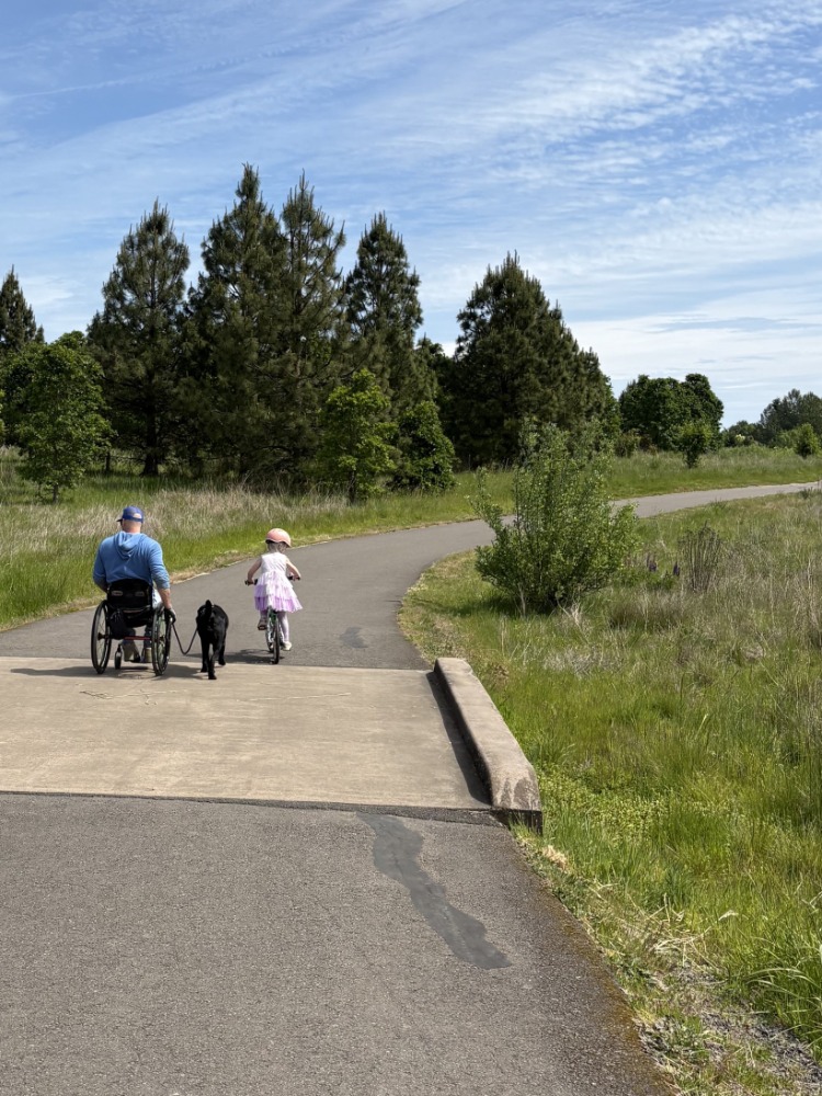 A person in a wheel chair on a cement trail next to a child on a bike.
