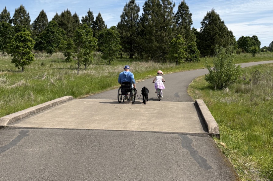 A person in a wheel chair on a cement trail next to a child on a bike.