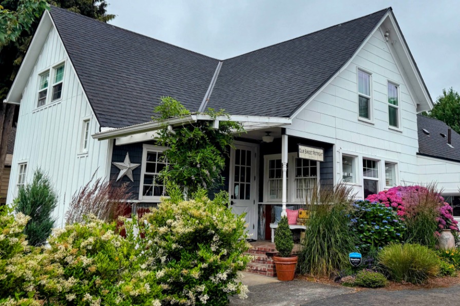 A white farm house style building with a black roof and green bushes out front