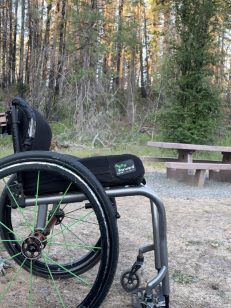 A wheelchair sits in the dirt outside a tent with a picnic table in the background