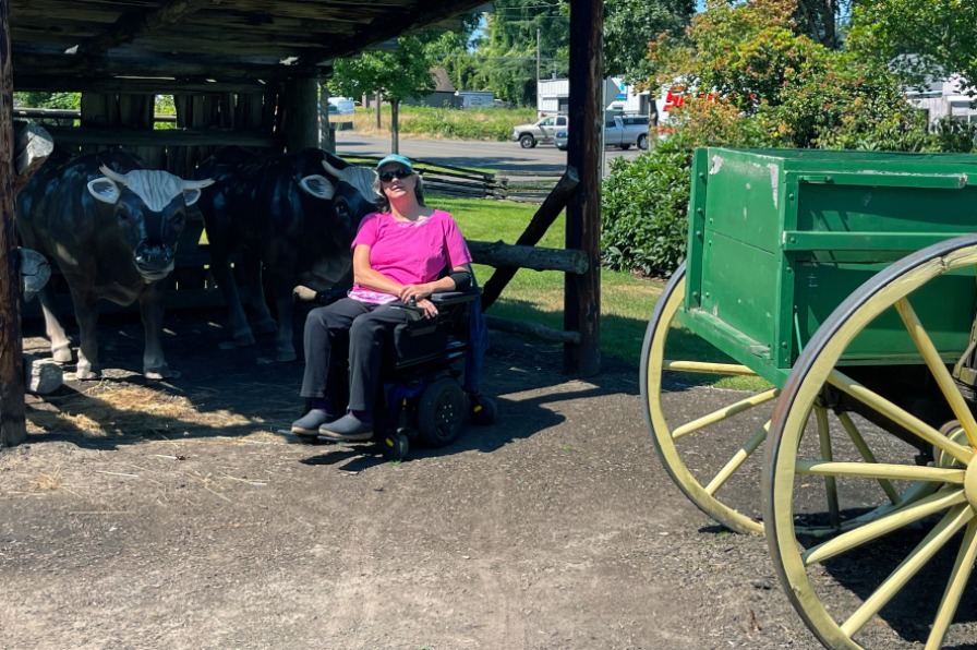 A visitor in a wheelchair poses outside with ceramic oxen and a wagon