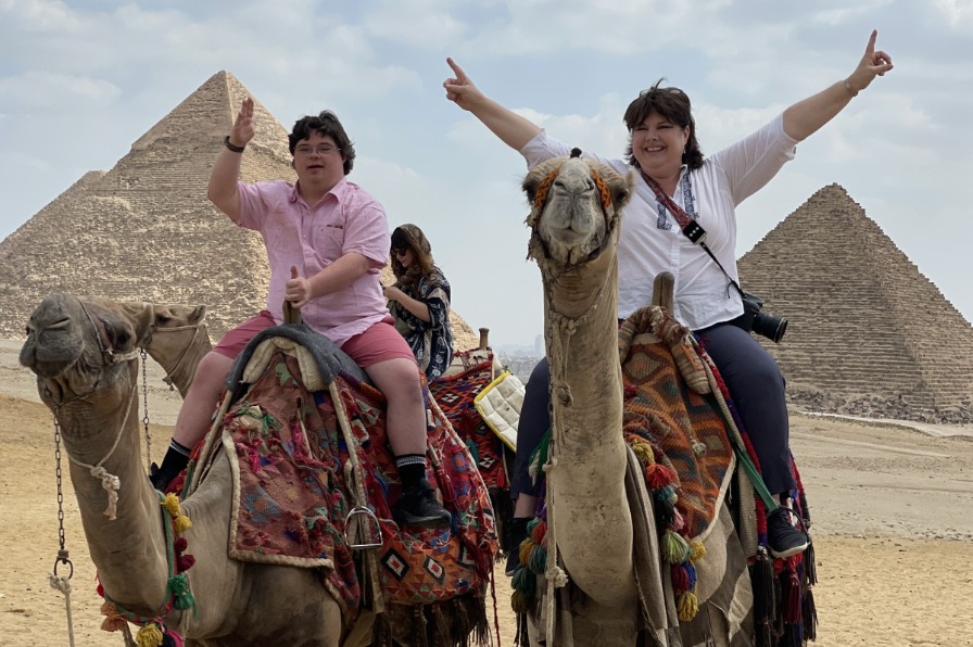 Downs & Towns Two people sit on two brown camels with tan pyramids in the back