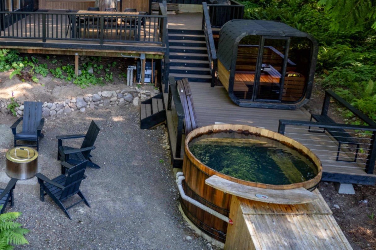 Black colored cabin tucked into the woods with an expansive deck featuring a sauna and hot tub. 