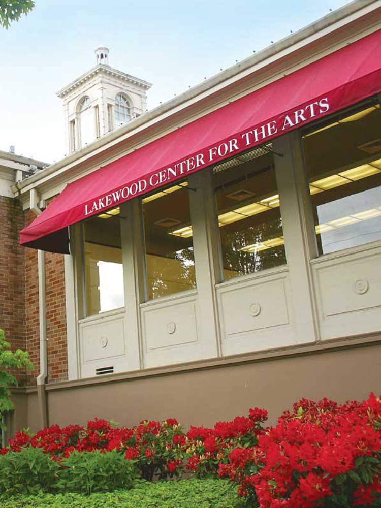 Exterior view of the Lakewood Center for the Arts, featuring large windows, red awnings, and vibrant flower beds. Lush greenery surrounds the building.