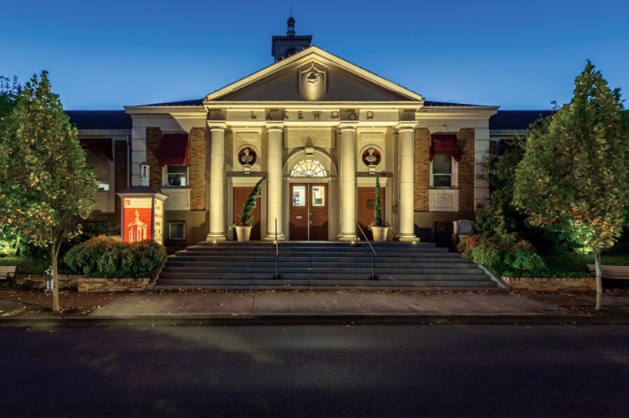 Historic building with grand columns and illuminated entrance, flanked by trees, set against a twilight sky. Steps lead to the main doors.