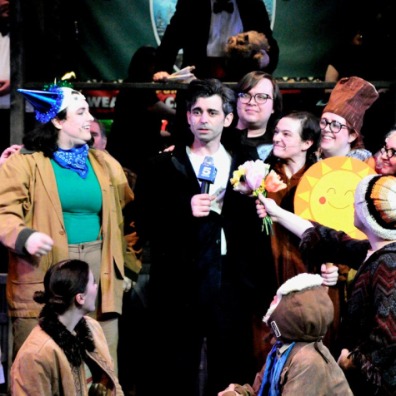 A lively stage scene featuring a group of performers celebrating, with a banner reading "Home of Punxsutawney Phil" in the background.