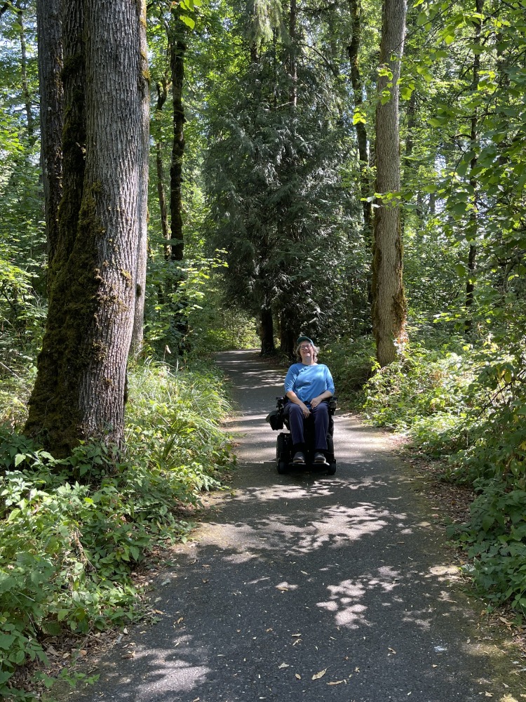 A wheelchair users in a blue shirt is on a paved trail surrounded by green trees.
