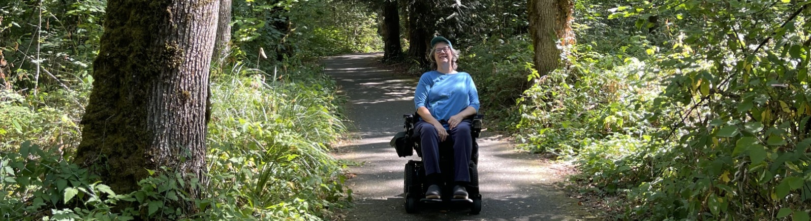 A wheelchair users in a blue shirt is on a paved trail surrounded by green trees.
