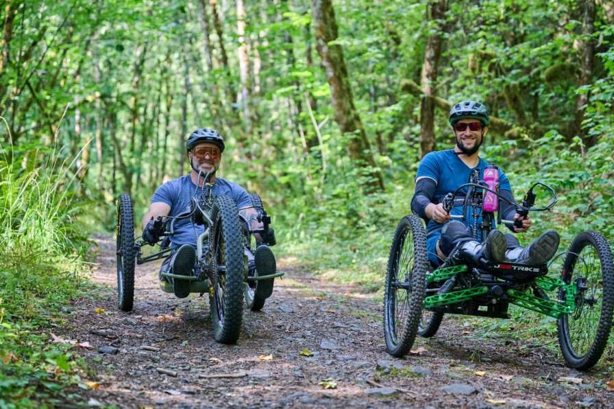 Two cyclists on adaptive mountain bikes on a dirt trail surrounded by green trees