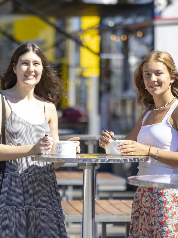 Two people in sundresses eat at tall tables surrounded by food carts
