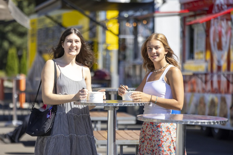 Two people in sundresses eat at tall tables surrounded by food carts