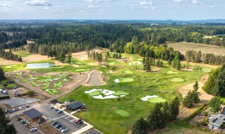 Aerial view of a golf course surrounded by lush trees and open fields, with a parking area and nearby buildings visible.