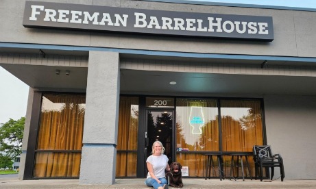 A person sits with a chocolate lab outside Freeman Barrel House, a casual establishment with large windows and warm curtains. The sign is prominently displayed above the entrance.