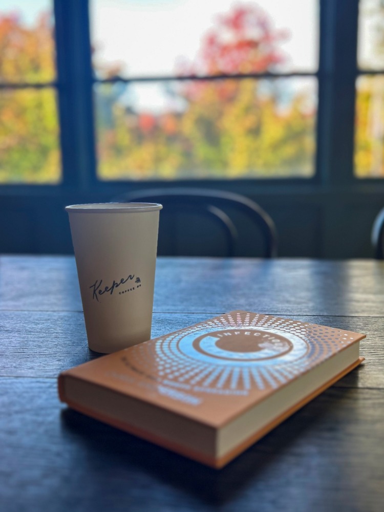 Cup of Keeper Coffee on a table next to a book with fall foliage in window outside