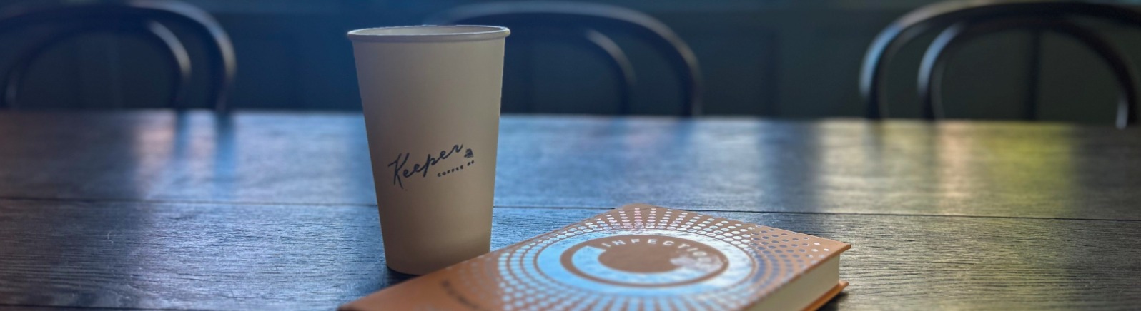 Cup of Keeper Coffee on a table next to a book with fall foliage in window outside