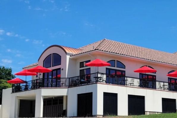 A modern building with a tiled roof features a spacious terrace adorned with red umbrellas, set against a clear blue sky.