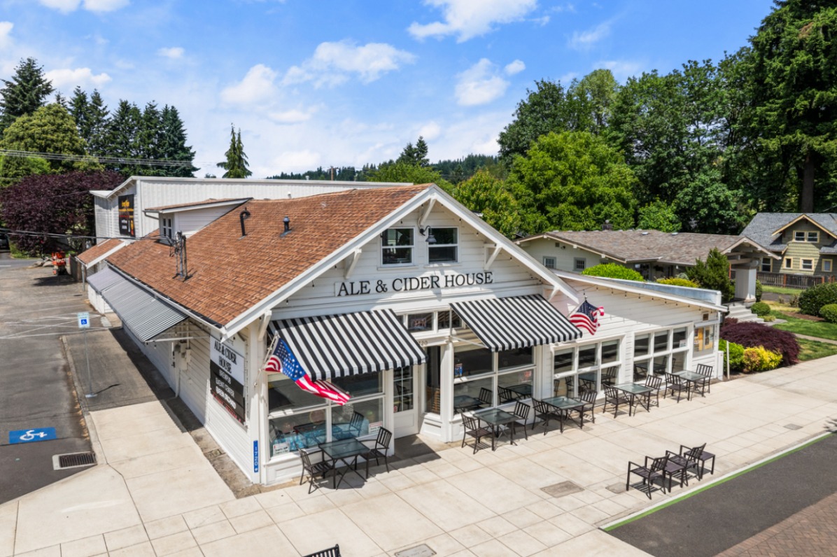 Ale and cider house with outdoor seating, featuring striped awnings and American flags, surrounded by greenery and blue skies.