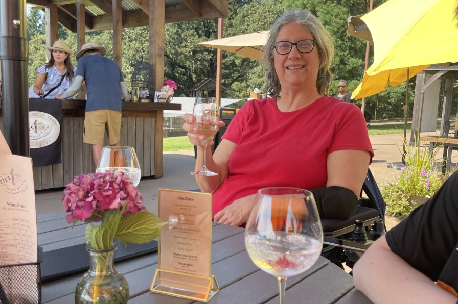 A visitor holds a glass of wine on an outdoor patio with green trees and yellow umbrellas behind.