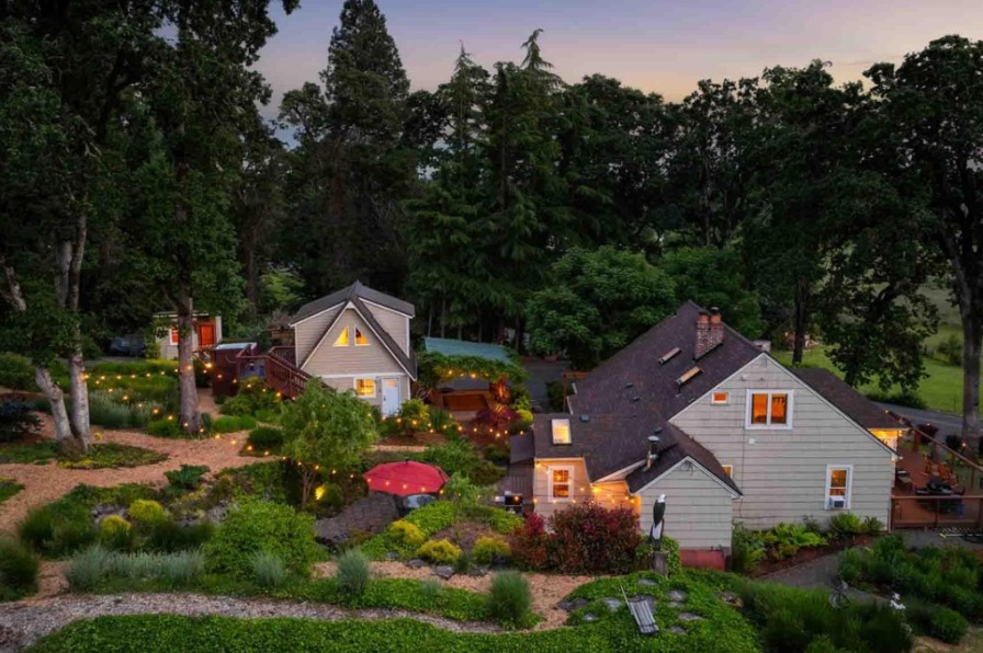Aerial view of a charming, illuminated home surrounded by lush gardens and trees at dusk, creating a serene and inviting atmosphere.