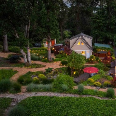 Aerial view of a charming, illuminated home surrounded by lush gardens and trees at dusk, creating a serene and inviting atmosphere.