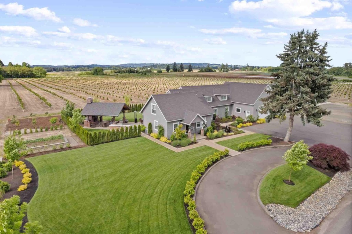 Aerial view of a charming gray house surrounded by lush green lawns and neatly arranged flower beds, with expansive orchards in the background.